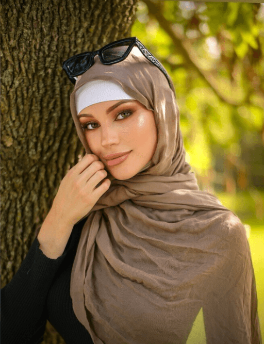 Portrait of a woman in a beige hijab with a white undercap, sunglasses on her head, posing beside a tree in a sunlit park.