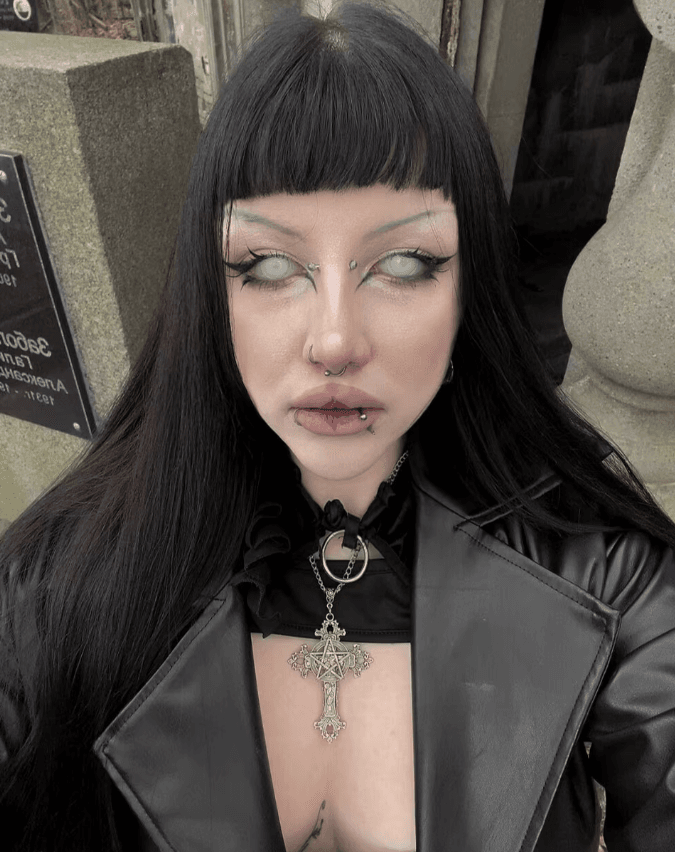 Person with long black hair and bangs, wearing a black leather jacket and a large ornate cross necklace, with pale makeup and facial piercings.