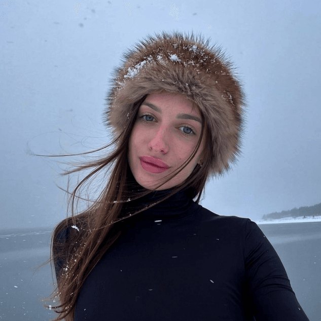 Woman wearing a fur-trimmed hooded coat outdoors in falling snow, facing the camera with windblown hair and blue skies behind.