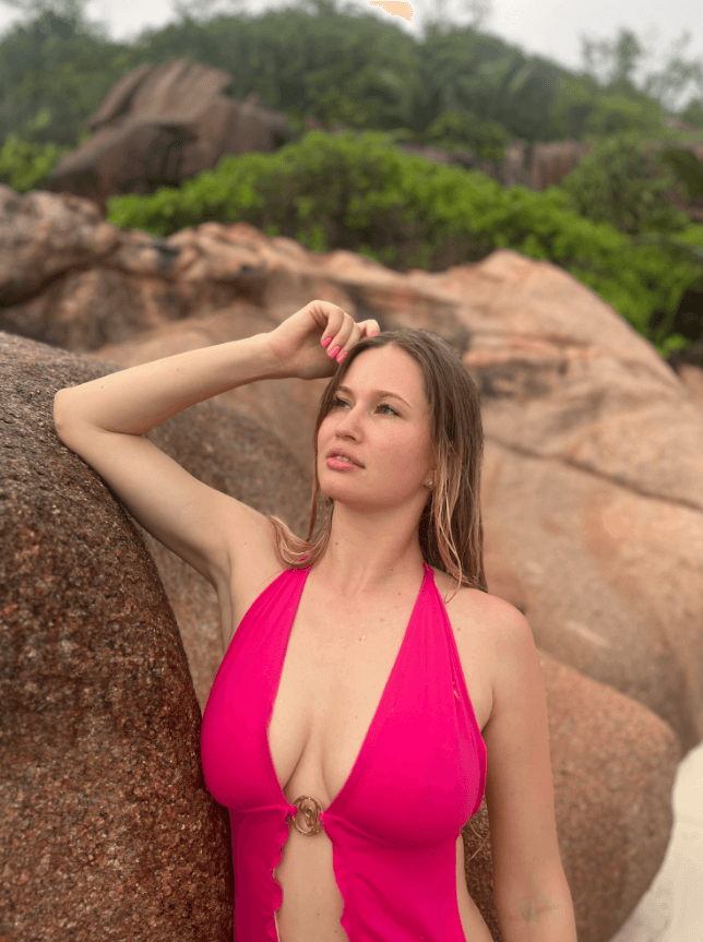 Woman in a pink halter swimsuit leaning against a rocky beach wall, looking into the distance.