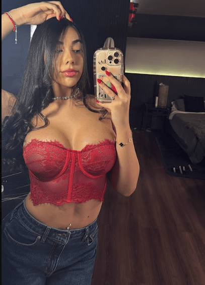Woman taking a mirror selfie in a red lace bustier top and jeans, holding a gold iPhone case and posing with one hand in her hair.