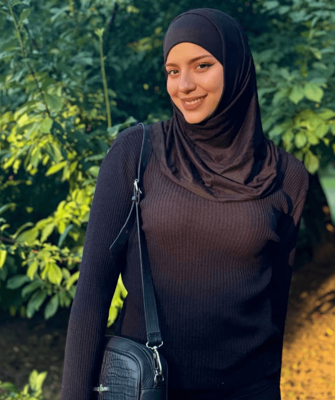 Woman wearing a black hijab and sweater smiling outdoors with greenery in the background.