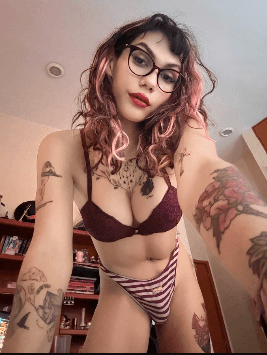 Selfie of a tattooed woman with pink hair and glasses wearing burgundy lingerie in a bedroom.